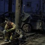 Un bombero triste y sentado en lo que son los restos de un auto incinerado a sus espaldas