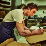 Un cocinero en medio de su lugar de trabajo escribiendo algo en un pedazo de carton