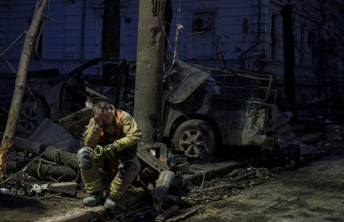 A sad firefighter sitting among the remains of a burned-out car behind him